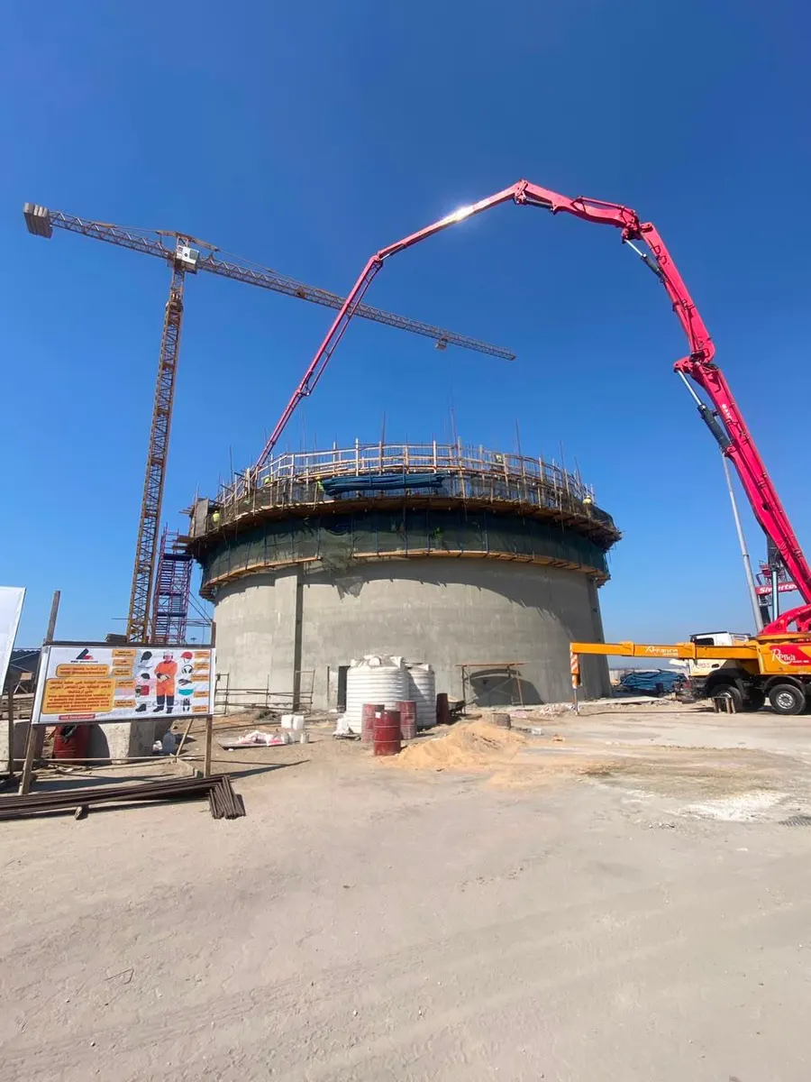 Concrete pump boom reaching over the top of a silo, with a tower crane and site safety signage alongside