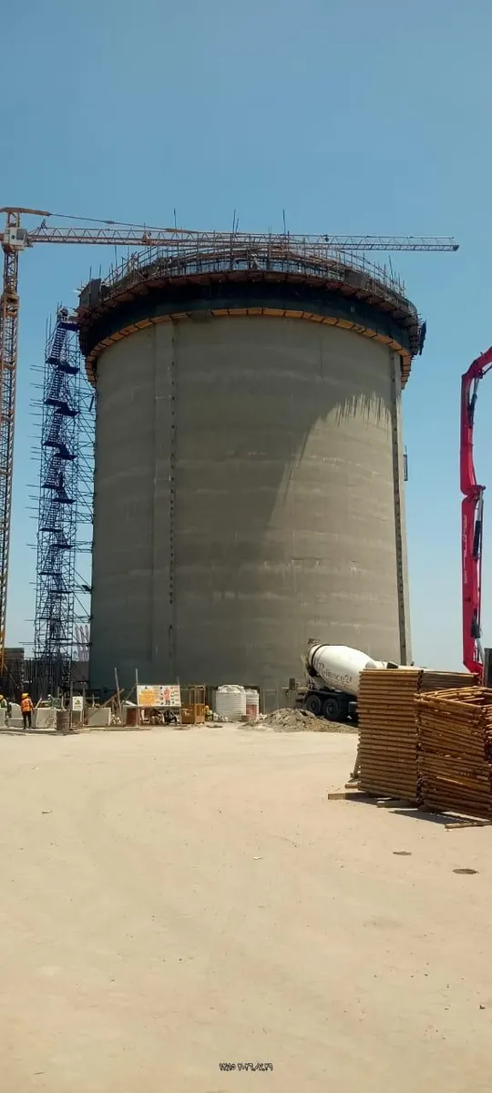 Concrete mixer truck at the base of a silo with stacked scaffold planks and a pump boom alongside