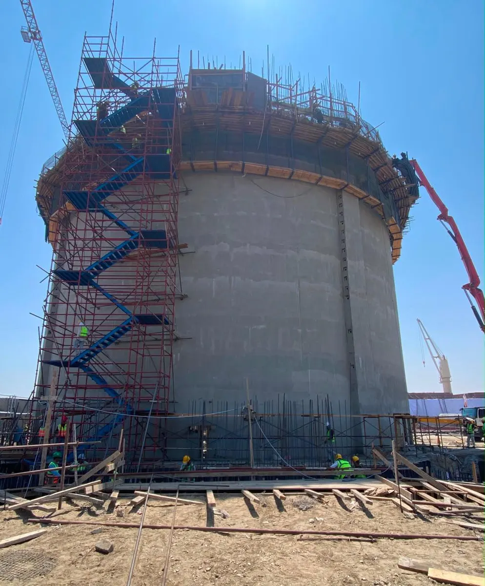Scaffold stair tower rising alongside a silo wall, with crews working on the base slab formwork below