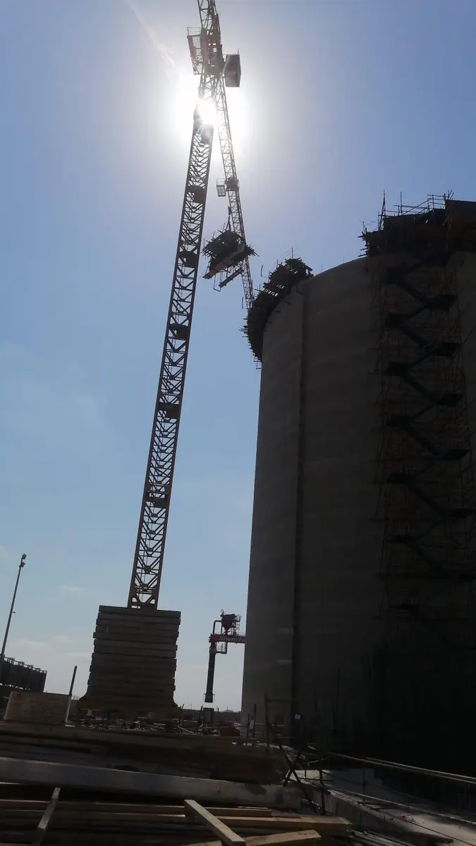 Tower crane silhouetted against the sun beside a scaffolded silo under construction