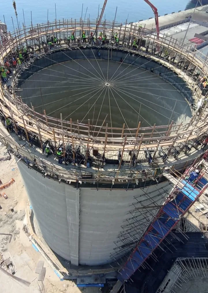 Crews working atop the cylindrical wall of a cement silo under construction at the Bulk Cement Export Station, with the quay and sea behind